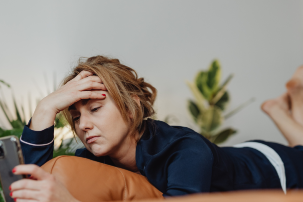 jonge vrouw ligt op de bank met mobiel in de hand en kijkt vermoeid