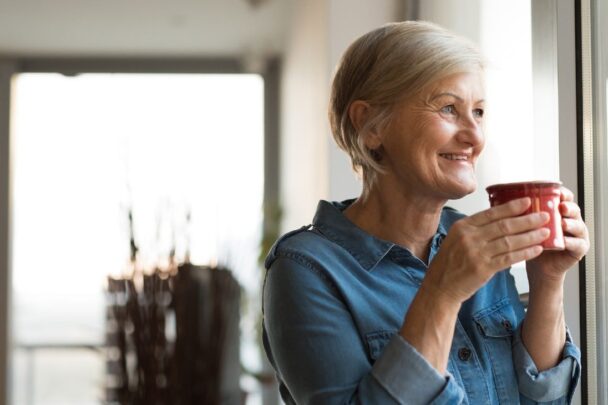 oudere vrouw kijkt lachend uit het raam en drinkt uit een mok