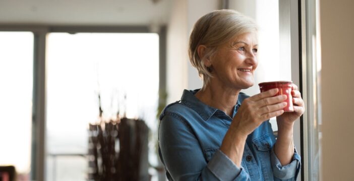 oudere vrouw kijkt lachend uit het raam en drinkt uit een mok