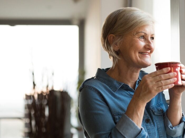 oudere vrouw kijkt lachend uit het raam en drinkt uit een mok