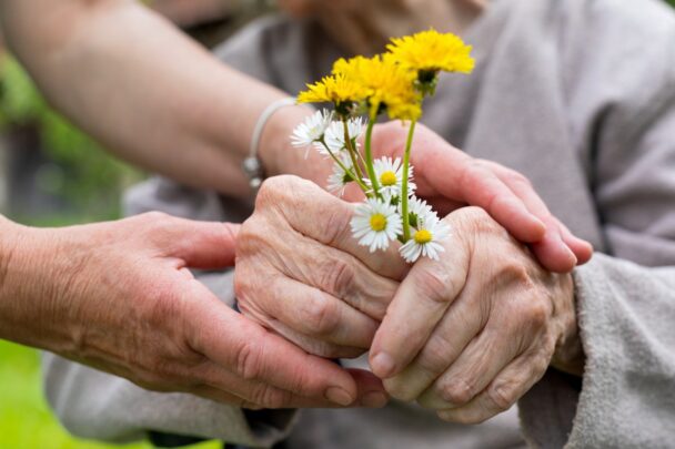 Oudere handen houden een bosje wilde bloemen vast. Daarbij iemand met jongere handen die ondersteunt.