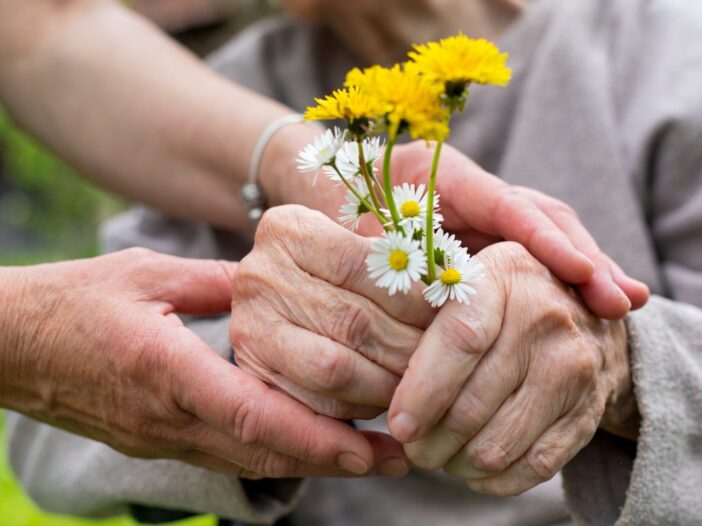 Oudere handen houden een bosje wilde bloemen vast. Daarbij iemand met jongere handen die ondersteunt.