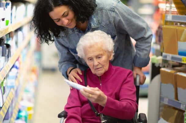 Oudere mevrouw zit in een rolstoel in de supermarkt. Jongere vrouw kijkt met haar mee op een stuk papier.