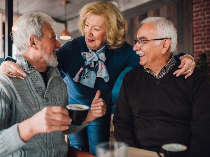 Twee oudere mannen drinken samen koffie en een oudere vrouw staat er gezellig bij.