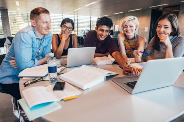 jonge mensen zitten aan een tafel met elkaar te lachen en zijn omringt met studie spullen zoals laptops en boeken.