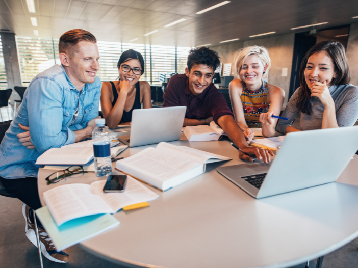 jonge mensen zitten aan een tafel met elkaar te lachen en zijn omringt met studie spullen zoals laptops en boeken.