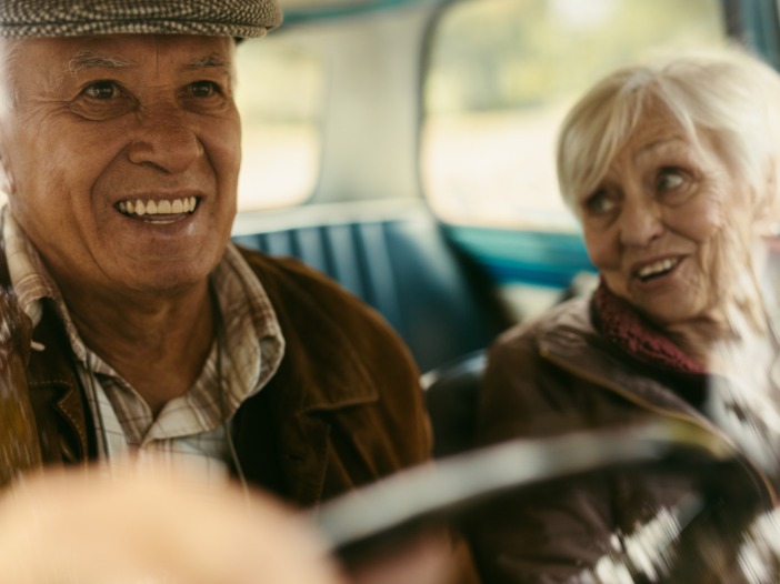 Twee ouderen mensen in een auto samen. Ze lachen heel leuk samen en de man brengt de vrouw naar haar bestemming.