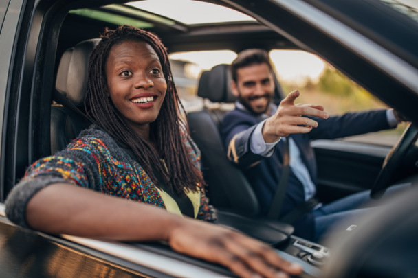 Twee lachende mensen samen in een auto. De man wijst ergens naar en de vrouw lacht, de man vervoert de vrouw.