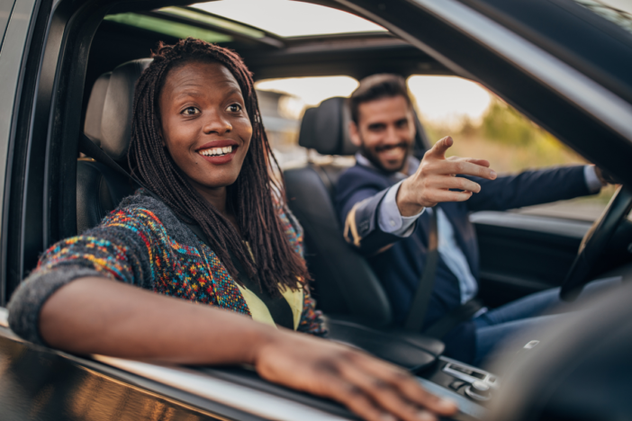 Twee lachende mensen samen in een auto. De man wijst ergens naar en de vrouw lacht, de man vervoert de vrouw.