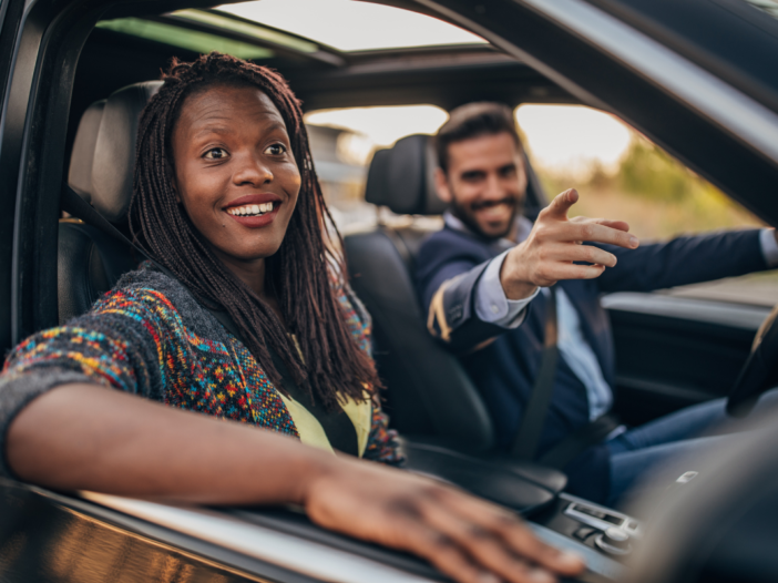 Twee lachende mensen samen in een auto. De man wijst ergens naar en de vrouw lacht, de man vervoert de vrouw.