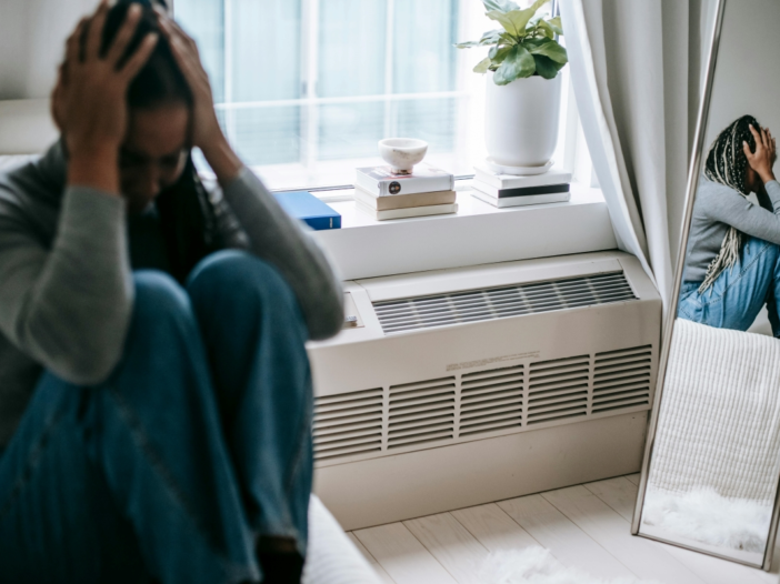 vrouw zit met haar handen in haar haar in huis. Ze maakt een verdrietige indruk.