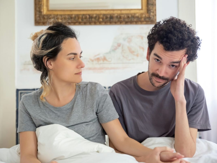 man en vrouw zitten samen rechtop in bed. Man ziet er gespannen of bezorgd uit.