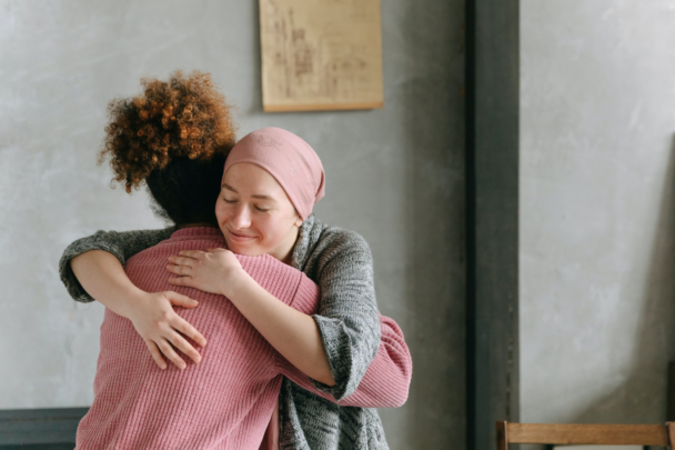 vrouw knuffelt een andere vrouw. Een van de twee oogt ziek en heeft een doek op haar hoofd die haarverlies verbergt.