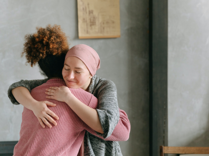 vrouw knuffelt een andere vrouw. Een van de twee oogt ziek en heeft een doek op haar hoofd die haarverlies verbergt.