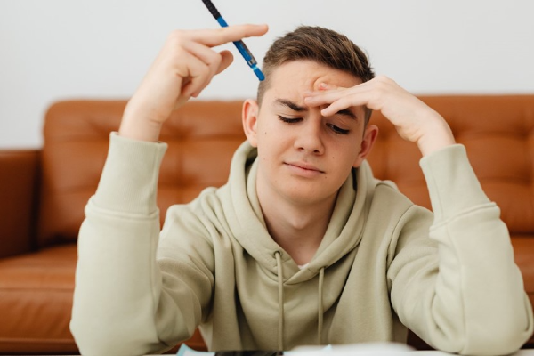 jongen kijkt met een moeilijke blik naar beneden, naar een stuk papier op tafel. Hij heeft een pen in zijn hand.