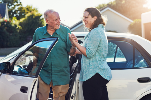 Vrouw helpt oude man uit de auto te stappen. De auto is wit en ze lachen allebei vriendelijk naar elkaar.