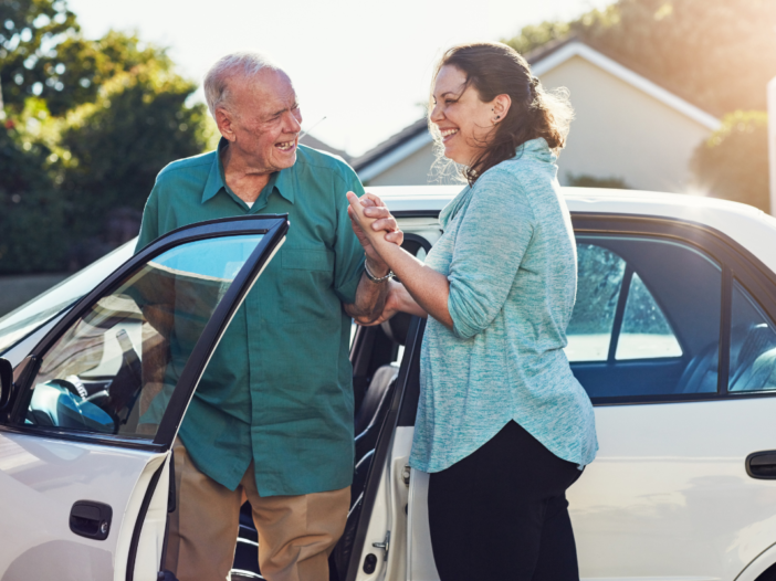 Vrouw helpt oude man uit de auto te stappen. De auto is wit en ze lachen allebei vriendelijk naar elkaar.