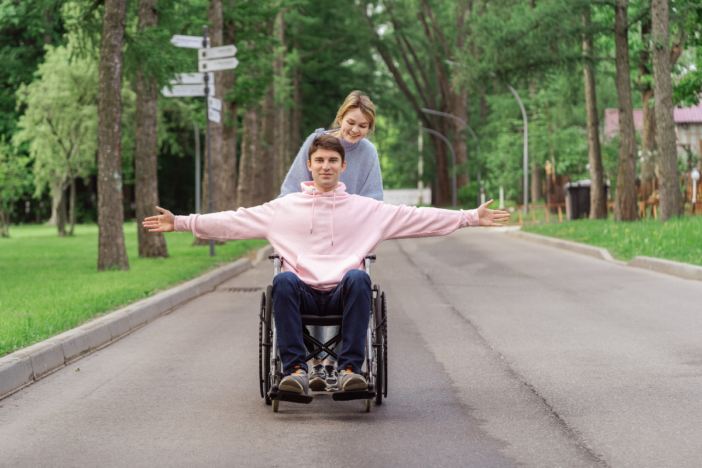 Jongen die in een rolstoel zit wordt geduwd door een meisje. Hij rijdt op de weg met armen gespreid onder de bomen.