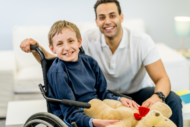 Kind met beperking in rolstoel. Man houdt zijn rolstoel vast. Kind heeft ook een knuffel in zijn handen.