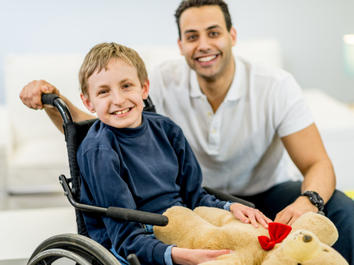 Kind met beperking in rolstoel. Man houdt zijn rolstoel vast. Kind heeft ook een knuffel in zijn handen.