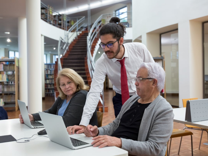 Man met een bril in een pak helpt twee oudere vrouwen met een laptop. Ze zitten in een bibliotheek.