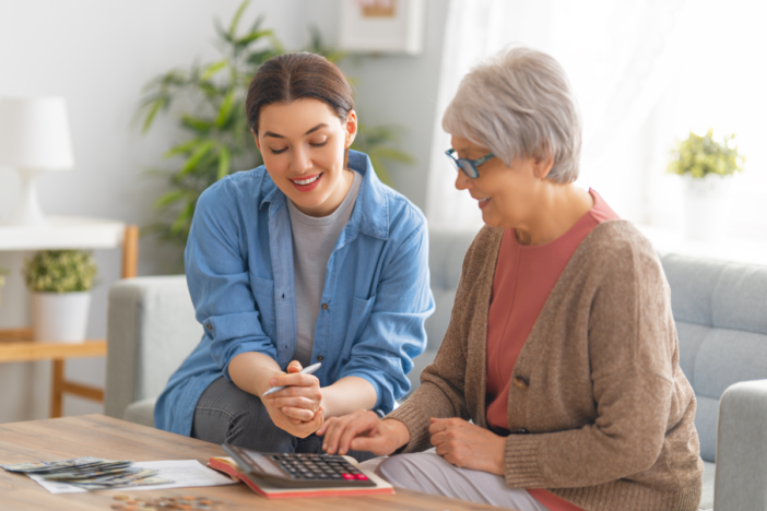 Vrouw helpt oudere vrouw met een rekenmachine. Ze zitten naast elkaar en lachen.