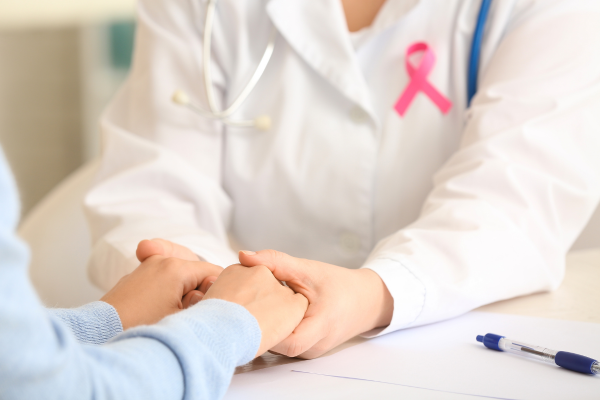 Dokter en patiënt zitten samen aan tafel en dokter houdt handen van patiënt vast. Dokter draagt een Pink Ribbon lintje