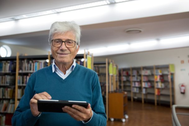 Man met grijze haren en een bril op lacht naar de camera. Hij is te vinden in de bibliotheek met een tablet in zijn handen om zo digitaler te worden,