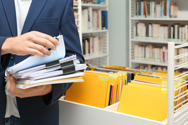 Man heeft mappen vast en staat bij de administratie. Hij heeft heel veel dossiers en gele mappen.