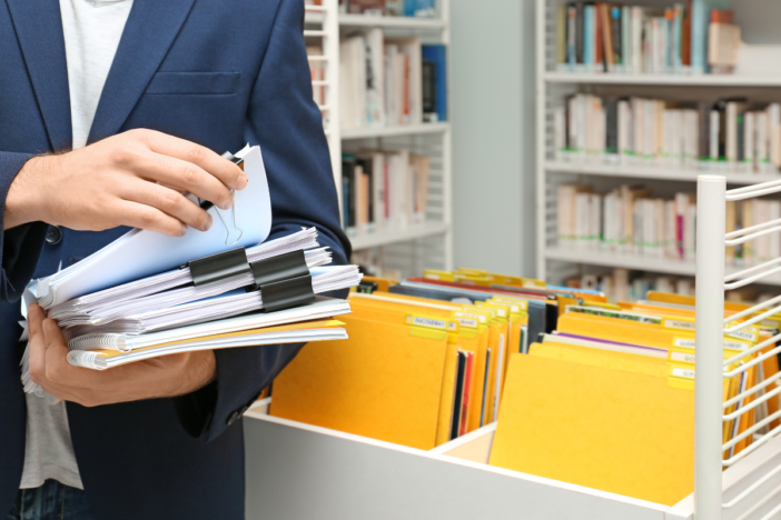 Man heeft mappen vast en staat bij de administratie. Hij heeft heel veel dossiers en gele mappen.