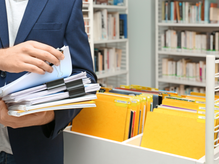 Man heeft mappen vast en staat bij de administratie. Hij heeft heel veel dossiers en gele mappen.