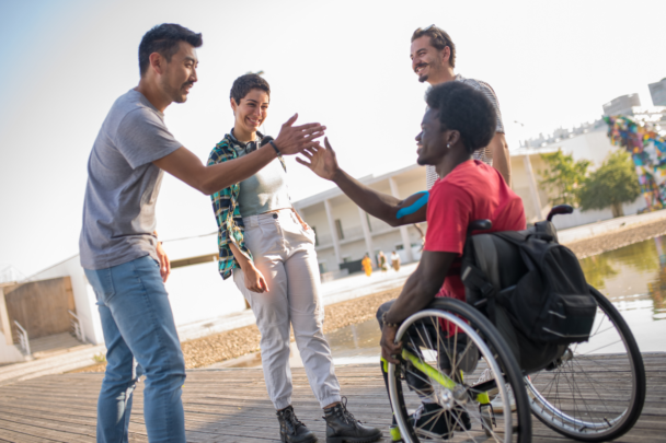 Een kind zit in een rolstoel en geeft een high-five aan een man. Ze zijn buiten, er staan nog twee mensen bij.