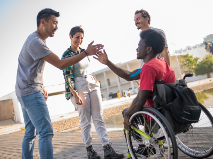 Een kind zit in een rolstoel en geeft een high-five aan een man. Ze zijn buiten, er staan nog twee mensen bij.