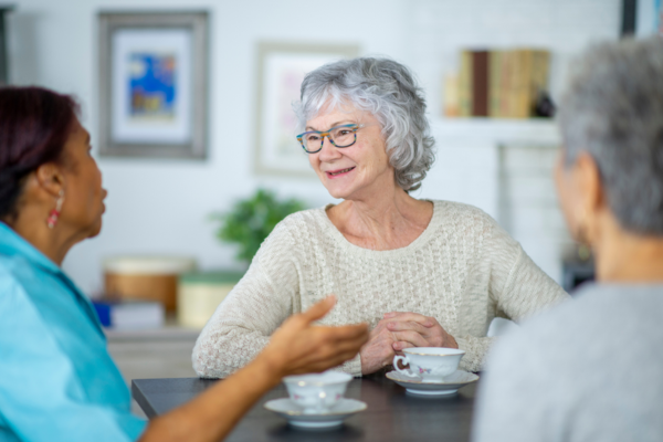 3 oudere dames zitten aan tafel in een kamer te praten. Op tafel staat koffie. Een dame lacht.