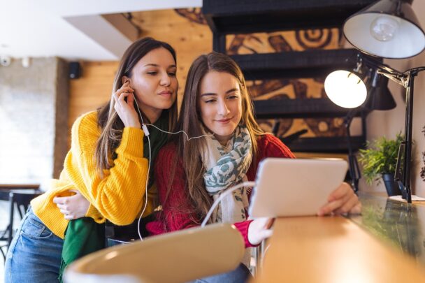 Twee tienermeiden luisteren samen via koptelefoonoortjes naar iets wat zich op een laptop afspeelt.