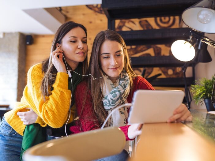 Twee tienermeiden luisteren samen via koptelefoonoortjes naar iets wat zich op een laptop afspeelt.