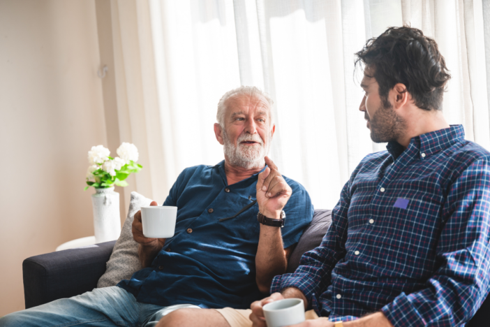 Twee mannen naast elkaar op de bank. Een senioren man en een volwassen man. Ze zijn aan het praten naar elkaar en de senior legt iets uit.