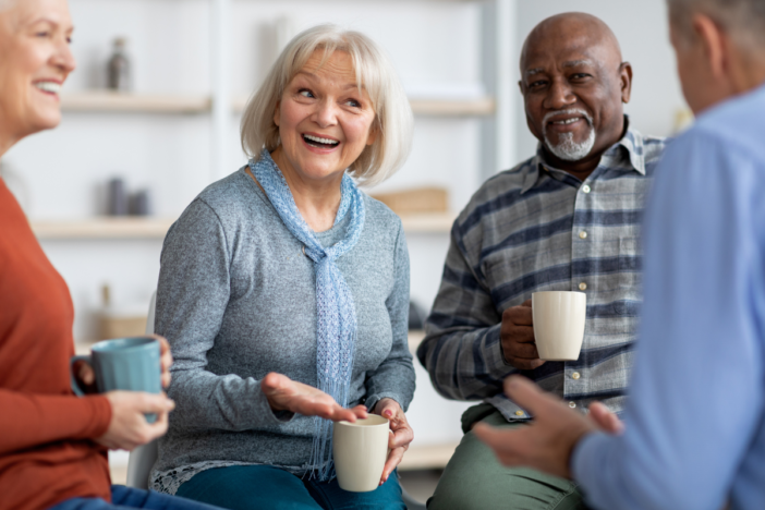 Vier senioren lachen samen en drinken een kop koffie. Ze zitten in een kring en de mevrouw in het midden is aan het lachen en aan het vertellen.