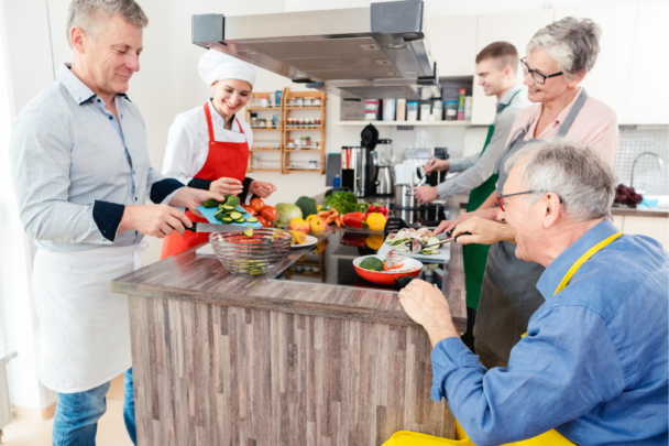 Vijf mensen die samen aan het koken zijn. Sommige mensen hebben dementie en sommige zijn er om te helpen en ondersteunen en vooral gezelligheid te maken.