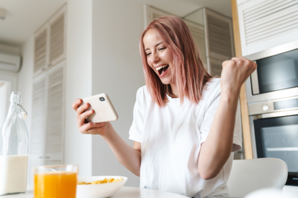 Vrouw kijkt naar haar telefoon en wordt helemaal blij. Ze zit aan de ontbijttafel en heeft net geld bespaard, daarom is ze zo blij.