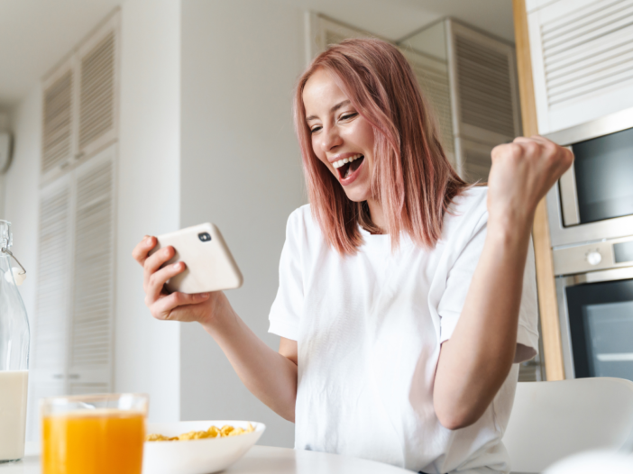 Vrouw kijkt naar haar telefoon en wordt helemaal blij. Ze zit aan de ontbijttafel en heeft net geld bespaard, daarom is ze zo blij.