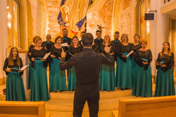 Een koor die in de kerk aan het zingen is. Ze hebben allemaal een groene rok aan en kijken naar de dirigent.