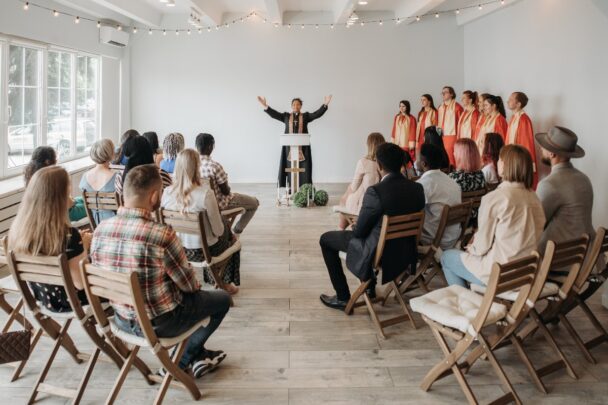 Een koor optreden in een kerk. Er zit publiek te kijken en het koor heeft een rood pak aan. De dirigente staat met haar handen omhoog en heeft een zwart pak aan met een ketting met een kruis.