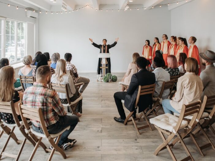 Een koor optreden in een kerk. Er zit publiek te kijken en het koor heeft een rood pak aan. De dirigente staat met haar handen omhoog en heeft een zwart pak aan met een ketting met een kruis.