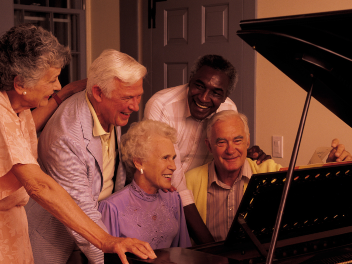 Groep oudere mensen die met z'n alle achter de piano staan. Witte mensen en donkere mensen achter de piano.