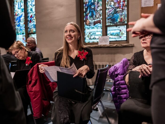 Lachende vrouw met notenboek in haar handen. Ze draagt een mooi zwart pak met een roos en ze zit in de kerk.