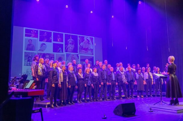 Impressie van het koor in Geldrop. Blauwe lichten en veel mensen op het podium die aan het zingen zijn.