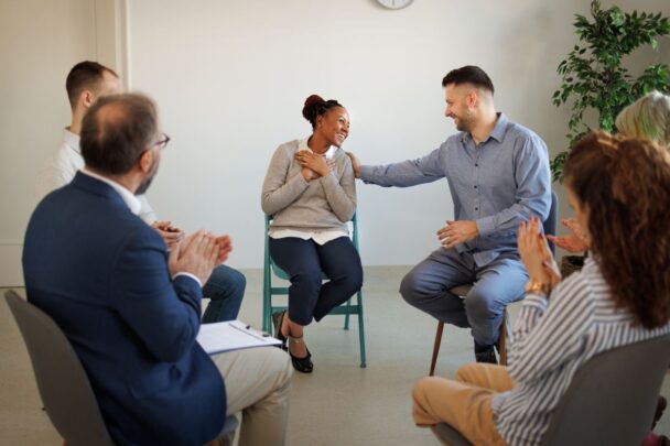 Mensen zitten in een kring en praten met elkaar. Man legt zijn hand op de schouder van een vrouw in de kring om medeleven te tonen.