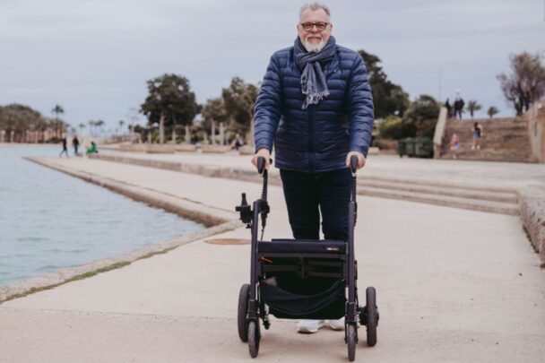 Een man staat buiten met een rollator. Hij heeft een jas en een sjaal aan.