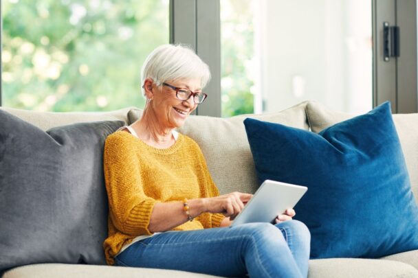 Mevrouw van oudere leeftijd zit op de bank. Ze klikt met haar vinger op het scherm van een ipad.